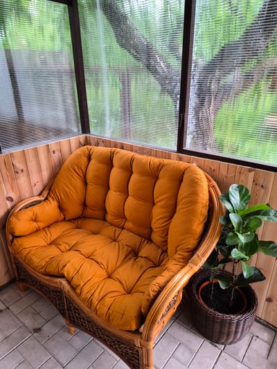 The image depicts an enclosed corner of a structure, possibly a veranda or sunroom, located in Hlevakha, Ukraine. The structure features wooden paneling on the lower walls and corrugated, translucent polycarbonate or plastic sheeting for the upper walls and windows. The exterior surface of the window panels shows numerous small water droplets, indicating recent precipitation or condensation. Through the ribbed windows, a blurred view of dense green foliage and tree trunks is discernible. The floor is covered with light grey square tiles arranged in a herringbone or basketweave pattern.

A two-seater sofa occupies the corner, featuring a woven rattan or wicker frame in a light brown hue. The sofa is fitted with thick, tufted cushions of a rust-orange or burnt orange color on both the seat and backrest. To the right of the sofa, a plant is situated on the floor. The plant, possibly a Ficus lyrata given its large, dark green leaves, is contained within a terracotta-colored pot, which is then placed inside a dark brown woven basket. There are no individuals or discernible actions present in the scene.