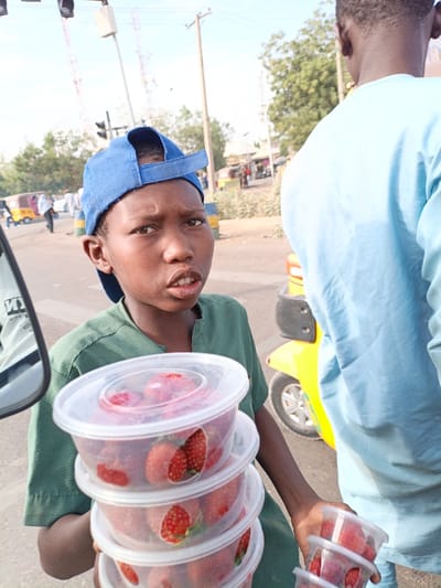 A young boy wearing a green shirt and blue baseball cap holds several clear plastic containers filled with strawberries. He looks directly at the camera with a slight frown. Another person stands to the right, partially obscuring the background. A yellow three-wheeled vehicle is visible behind them. The scene is outdoors on a sunny day, with traffic lights and other people in the blurred background.