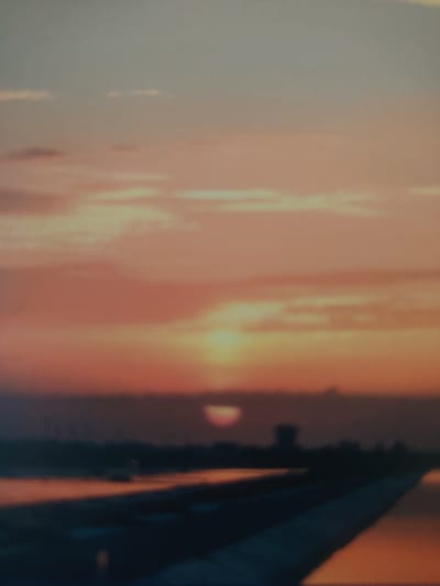 The image captures a hazy sunset over a body of water, likely a canal or river, with a distant shoreline visible. The sky is dominated by warm hues of orange, pink, and pale blue, with soft clouds. A dim, partially obscured sun is setting near the horizon, casting a reddish glow. In the middle ground, a dark silhouette of what appears to be a building or structure, possibly a tower, stands against the sky. The foreground shows a dark, indistinct area that could be land or a path, with reflections of the sunset on the water's surface. The scene suggests a waterfront area within a city, but the specific location in Bahshitpa, Uzbekistan, cannot be determined from the visual information alone. There are no visible people or clear actions taking place.