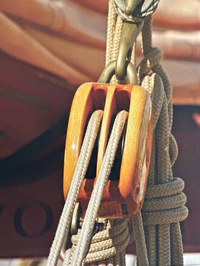 This is a close-up, eye-level shot of a wooden block and tackle system with thick rope, likely from a sailing vessel. The block is made of polished, varnished wood and has two sheeves where the rope runs. The rope is a thick, beige, braided material. A metal hook is attached to the top of the block, connecting it to a larger rope that is looped around it. In the background, out of focus, is a reddish-brown surface that appears to be part of a boat or sail, with some diagonal lines suggesting folds or structure. The lighting is bright, casting subtle shadows and highlighting the textures of the wood and rope. The overall impression is one of maritime equipment in use or being stored. The image does not contain any elements that definitively place it in Ossenzijl, Netherlands.