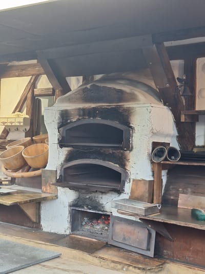 This image depicts an outdoor pizza oven with a rustic, old-world aesthetic. The oven is constructed of white stucco, showing signs of age and soot accumulation around its openings. It has two distinct oven chambers stacked vertically, with the lower one exhibiting glowing embers, indicating it is currently in use. Above these chambers, the oven is topped with a curved, black dome.
To the left of the oven, a collection of five large, wooden bowls are arranged on a curved wooden rack, suggesting they are used for dough or serving. Behind the bowls, a small decorative wall-mounted unit with three drawers and hooks is visible.
The oven and its surroundings are supported by wooden beams and framing, contributing to the rustic feel. To the right of the oven, a metal shelf holds two dark, cylindrical objects. Further to the right, a wooden counter surface is visible, with a metal pan and utensils resting on it, as well as a green brush. A metal door lies on a wooden surface in the foreground. The overall environment appears to be an outdoor seating or preparation area, possibly part of a restaurant or a communal gathering space. The lighting suggests it is daytime, with the sun casting shadows.
This scene is located in Leipzig, Germany.