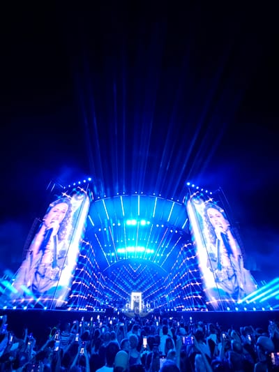 The image depicts a concert at PGE Narodowy stadium in Warsaw, Poland. The stage is illuminated with bright blue and white lights, featuring large screens displaying a female performer. A crowd of concertgoers is visible in the foreground, many holding up their phones to capture the event. The overall atmosphere is that of a lively outdoor music performance.