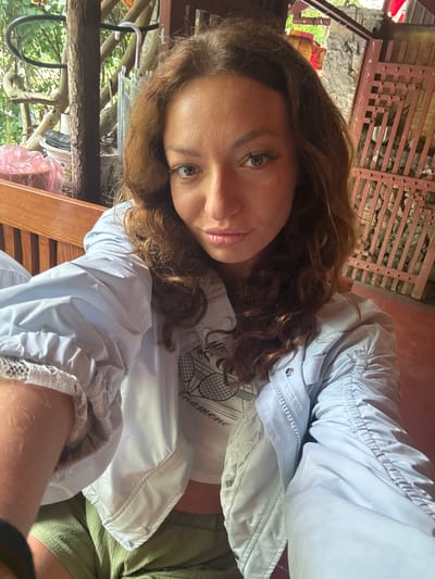 A woman poses indoors in Mèo Vạc Commune, Vietnam. She has brown curly hair and light skin. She wears a light blue jacket over a white t-shirt and green bottoms. The setting includes a rustic wooden structure, likely a building, with a reddish-brown lattice wall in the background. Other wooden elements, possibly a fence or structure, are also visible with glimpses of greenery. The lighting is soft and natural.