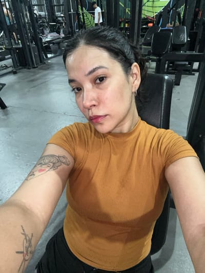 The image depicts a woman taking a selfie inside a gym. She is positioned in the foreground, facing the camera, with her torso and head visible. She has dark hair pulled back, light brown skin, and is wearing a mustard yellow, short-sleeved, ribbed t-shirt. Small hoop earrings are visible in her ears. Her left arm displays two tattoos: one of a stylized female face with a bun on her bicep, and a second, partially visible tattoo of a hand holding an object on her forearm. Her skin appears slightly dewy, consistent with physical exertion.
She is seated on a black padded bench, part of a piece of gym equipment. The background reveals a fitness environment with multiple black metal exercise machines, weight racks, and benches. The floor is light gray concrete. Two other individuals are faintly visible in the mid-ground: one person in a white t-shirt stands near equipment, and another is partially obscured further back. A green wall element, possibly part of a mural or logo, is visible behind some equipment. The lighting is artificial.
The specific location within Ciudad del Libertador General San Martín, Argentina, cannot be determined from the visual information provided; the image only indicates a gym setting.