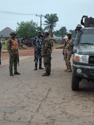The image shows a group of soldiers in Gidan Waya, Nigeria, standing at attention on a paved street. The soldiers are wearing camouflage uniforms and black boots. One soldier is holding a rifle. A military vehicle is parked nearby. The environment consists of trees, houses, and a utility pole. The sky is overcast.