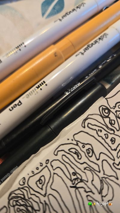 This close-up image captures various art supplies alongside a piece of paper displaying an abstract drawing. Several pens or markers are arranged diagonally across the frame. On the left, two white pens are visible, both branded with "kingart™". An orange marker lies between them and a black pen. The black pen, which appears to be a fineliner, features "marvU®" on its clip and "InkLine™ Pen" printed on its body. Below the black pen, partially visible text reads "Water-Based" on what seems to be its packaging or an adjacent white surface. These art tools rest on a patterned fabric, possibly a tablecloth, which displays a faint blue leaf design on the far left.
A white piece of paper occupies the right side of the frame, showcasing an intricate black line drawing composed of organic, interconnected shapes and loops, resembling a complex doodle or part of a larger artwork. In the top right corner of the image, a social media-style overlay adds a collection of emojis: a snail (🐌), a camera (📸), a face emoticon (•_•) , a sparkling star (✨), and a sprout (🪴). The scene suggests an ongoing or recently completed artistic activity, likely indoors on a table or desk, under soft, ambient lighting. No people are visible.