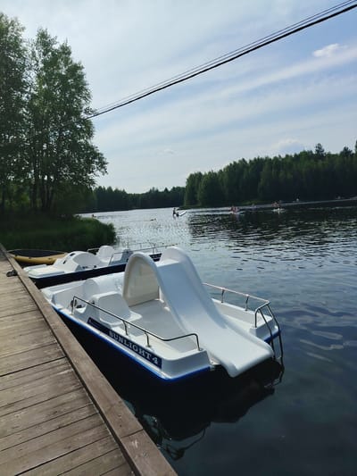 The image depicts a waterfront scene with several pedal boats docked at a wooden pier. The pedal boats are white with blue trim and one is labeled "SUNLIGHT 4". In the background, a lake is visible, with a dense forest lining the opposite shore. Several people are engaged in water activities on the lake, including paddling and what appears to be stand-up paddleboarding. The sky is partly cloudy, with power lines stretching across the upper portion of the frame. The scene suggests a recreational area, likely within the city of Vylytdor, Russia.