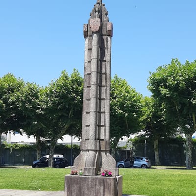 The image shows a tall, gray, stone monument in the city of Vila Verde, Portugal. The monument has a rough, textured surface and is made of stacked, irregular blocks, with a shield-like emblem near the top. The monument is located on a grassy area, with a sidewalk or paved path in the foreground. Behind the monument, there are several large, leafy green trees. In the background, there are cars parked on the side of the road, and a building with a dark, reflective facade. The sky is clear and blue. There are no people visible in the image.