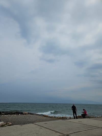 This vertical, eye-level, outdoor shot captures a portion of the coast in Güzelbahçe, Turkey. The scene is dominated by a vast, cloudy sky in muted blues and grays, with patches of lighter clouds suggesting an overcast day. Below the sky, the choppy, dark green sea stretches horizontally across the frame, meeting a rocky and pebbled shoreline. In the foreground, a concrete area with visible seams forms a walkway or promenade.

On the rocky area overlooking the sea, two individuals are present. A man, dressed in a black t-shirt and pants, stands facing the water, his back partially to the camera. To his right, a woman sits on a rock, wearing a pink top and a dark skirt. She is looking down, possibly at a device in her hands. The sea is active with visible waves and whitecaps. In the distance, a hazy line of mountains can be seen against the horizon. The overall atmosphere is calm but possibly indicative of approaching weather.