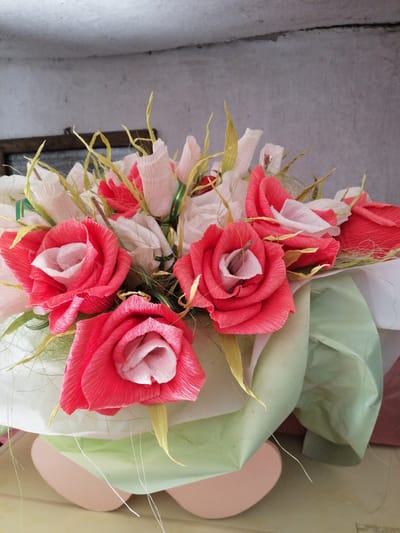 A close-up, eye-level, indoor shot displays a bouquet of artificial flowers, likely made from crepe paper, set against a textured, off-white wall. The bouquet features several prominent pink and white roses, interspersed with white buds and yellow decorative elements resembling grass or leaves. The flowers are arranged in a light green wrapping, with a white layer beneath. The background wall has a visible seam running horizontally across it. The base of the bouquet rests on a light-colored surface. The scene appears to be a still life, with no people or animals present. The image is sharp and well-lit, with a shallow depth of field that blurs the background. No specific location in Lanivtsi, Ukraine is identifiable from the image content.