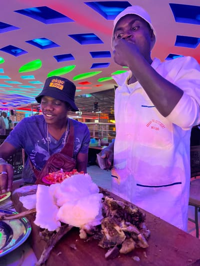 The image depicts two men sharing a meal at an eatery in Kimorori, Kenya, likely during the evening or night, given the artificial lighting. The scene is bathed in a purple and pink ambient glow from unseen light sources, complemented by a decorative wall featuring illuminated blue, green, and red geometric shapes.
One man, positioned on the left, is wearing a white uniform that resembles a chef's coat or a butcher's apron. On the left breast pocket of his uniform, red text is visible, which reads "ENDAI BUTCHERY MOISBO" (or MOISO). He looks directly at the camera with a neutral to slightly smiling expression, his right hand raised towards his mouth.
The second man, seated on the right, wears a blue t-shirt and a black bucket hat with yellow text that appears to say "GIKARA FIYU". He is smiling softly and looking down towards the food. A brown shoulder bag is visible across his chest.
The meal is laid out on a large wooden serving board. It features substantial mounds of a white, starchy food, characteristic of ugali, alongside a generous portion of chopped grilled meat, known as nyama choma. A small bowl containing a red or orange salad or salsa is also present, and a metal plate or bowl is partially visible. The overall atmosphere suggests a casual dining experience.
