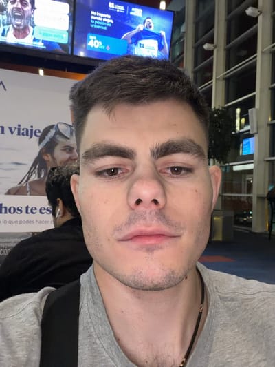 In a selfie taken at Aeropuerto Internacional Ezeiza, Argentina, a young man with short, dark hair and prominent eyebrows is shown from the chest up. He is wearing a gray t-shirt and a dark strap across his chest. His skin is light with some stubble on his chin and upper lip. His right eye is visible, appearing brown. In the background, a large advertisement features a woman wearing goggles and a bathing suit, with text in Spanish. Further in the background, a television screen displays an advertisement with a man celebrating and text in Spanish, alongside a "40% OFF" graphic. Another person wearing white earbuds is visible in the mid-ground, facing away from the camera. The environment appears to be an indoor public space with large windows and architectural elements typical of an airport.