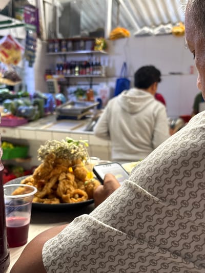 An indoor scene in Jesús María, Peru, captures a close-up perspective of a person engaging with food, likely within a market or a casual dining establishment. In the foreground, the upper torso and arm of a person are partially visible, wearing a light-colored garment adorned with a repeating pattern of intertwined "MK" logos, indicative of the Michael Kors brand. This individual's skin tone is medium to dark.

A black dish in the mid-ground holds a substantial portion of golden-brown fried food, appearing to be calamari rings or chicharrón de mariscos, garnished with fresh green herbs or small white blossoms. Adjacent to the food, a clear plastic cup contains a reddish-purple drink, consistent with chicha morada, a popular Peruvian beverage. A dark bottle holding a similar red liquid is also partially visible at the bottom of the frame.

The blurred background reveals another person in a white hooded sweatshirt, seen from behind. The setting is characterized by shelves stocked with various products, bottles, and packaged goods, suggesting a retail or food service environment. Additional background elements include what appear to be display racks showcasing small, golden-fried pastries or snacks. The scene depicts a casual meal being enjoyed in a busy, everyday setting.