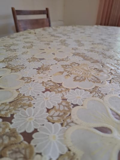 The image shows a close-up of a round tablecloth with a floral pattern. The tablecloth is predominantly white and gold and covers a table. In the background, a wooden chair with a slatted back is visible, along with a pale wall and a portion of a pink curtain. The scene appears to be indoors. There are no people or actions depicted in the image. The location is stated to be in the city of Galabovo, Bulgaria.
