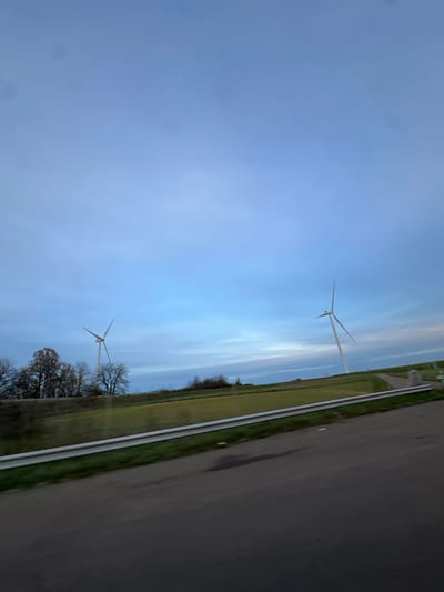 The image captures a panoramic view of a rural landscape near Sainte-Colombe, France, under a twilight or dawn sky. The upper two-thirds of the frame are dominated by a cool blue-grey sky with scattered clouds, exhibiting several dark, circular lens or sensor artifacts. The bottom third features a dark asphalt road.
Separating the road from the mid-ground is a long, silver metal guardrail running horizontally. Beyond the guardrail, a gently sloping green field extends across the mid-ground, showing patches of light and dark green vegetation. To the left, a cluster of deciduous trees, largely bare of leaves, forms a dark line against the horizon.
Two white wind turbines are the primary subjects. One turbine stands on the left, partially obscured by the trees. A second, more prominent turbine is positioned on the right, rising from the green field. Both turbines feature three blades, angled as if stationary or slowly rotating. To the right of the second turbine, a lighter-colored dirt path or access road ascends the grassy incline. Near the base of this path, indistinct low-lying structures or markers are visible. No individuals are present in the scene.