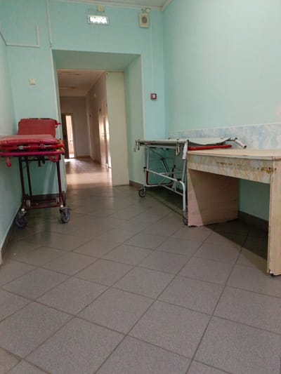 This is a ground-level, eye-level, medium-shot, indoor photograph taken in a hallway of what appears to be a medical facility in Zalesovo, Russia. The hallway is lit by artificial light, casting shadows on the tiled floor. The walls are painted a pale green.
In the foreground, to the left, is a red medical gurney with a padded mattress and wheels. To the right of the gurney is a white, metal medical trolley with wheels. Further down the hallway, there is a wooden desk with a worn surface.
The hallway opens into a further section of the building, where several closed doors are visible. Above the entrance to this section, an illuminated sign with Cyrillic text, likely "ВЫХОД" (Exit), is mounted on the wall. A small white speaker is also visible on the wall near the sign. To the left of the sign, a white electrical conduit runs vertically down the wall. A red fire alarm box is mounted on the wall to the left of the hallway.
The scene appears deserted. No people are visible in the image. The overall impression is one of a functional, perhaps dated, institutional environment.