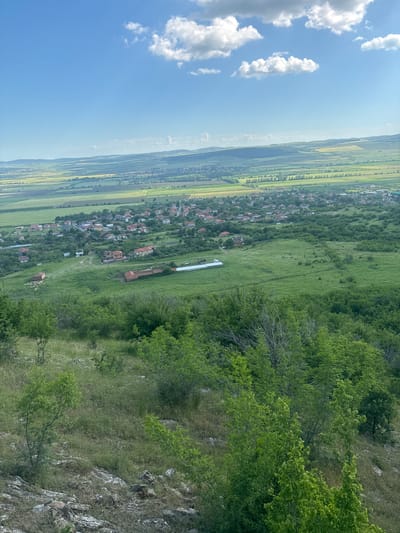 The image shows an aerial view of Goritsa, Bulgaria. The sky is blue with scattered white clouds. A patchwork of agricultural fields is visible, interspersed with a line of trees and the edge of a village. In the middle ground, red-roofed buildings and a long white structure are situated amidst green fields. The foreground is dominated by lush green trees and a rocky outcrop, suggesting a hillside vantage point.