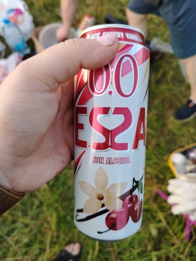 A hand is holding a can of "0.0 ES2A" flavored "Black Cherry Vanilla" non-alcoholic beverage. The can has a white background with red and yellow accents, and features graphics of a vanilla flower and cherries. The background is out of focus and shows green grass and the legs of at least two people. One person is wearing dark shorts and dark shoes, and the other is wearing dark sandals. There are also some plastic bags and a white bucket visible in the background. The scene appears to be outdoors, possibly at a picnic or gathering. There is no indication of the location in the city of Kriukovskoe, Russia.