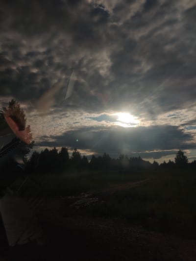 A landscape photograph captured in the city of Kamennyi Kliuch, Russia, depicts a scene under a heavily overcast sky with sunlight breaking through. The sun, positioned in the upper-middle right quadrant, emits intense crepuscular rays that penetrate dense, dark grey cloud formations. Patches of lighter sky are discernible near the sun's immediate area and intermittently across the upper frame.

A dark, silhouetted treeline, composed of numerous trees, forms a horizontal band across the middle of the image, separating the illuminated sky from the darker foreground. Below the treeline, an unpaved dirt track or road is visible, winding towards the right. Patches of standing water or mud are present on the track, and sparse, dark green vegetation flanks the road.

The image appears to have been captured from inside a vehicle, indicated by significant reflections on what is presumably a windshield. On the far left, a reddish-brown, indistinct reflection, possibly an ear or part of a head, is visible. Lighter, elongated reflections, indicative of windshield glare, extend into the upper left quadrant of the sky, and another distinct bright, angular reflection cuts across the upper right sky. No individuals or animals are discernible in the scene. The lighting is characterized by high contrast, with the bright sun creating deep shadows in the foreground and strongly silhouetting the trees.