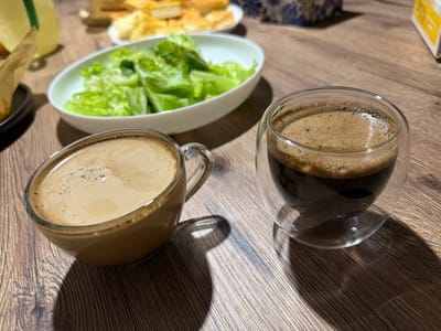 The image captures a close-up view of a wooden table set with two cups of coffee and a bowl of lettuce. The coffee on the left is in a ceramic mug with a handle, filled with a light brown, frothy liquid, suggesting it might be a latte or cappuccino. The coffee on the right is in a clear, double-walled glass cup, containing a darker, richer coffee with a thick crema, likely an espresso or Americano. Behind the coffee cups, a white oval bowl holds a portion of fresh, green lettuce leaves. Further in the background, out of focus, are glimpses of other food items on plates, including what appear to be pastries or bread, and a decorative object with a blue and white pattern. The lighting suggests it could be daytime, with shadows cast on the table from the cups. The setting appears to be a home or a cafe, with a cozy and relaxed atmosphere. No people are visible in the frame. The visible text is limited to a small yellow and white box in the upper right corner, which is too blurred to read.