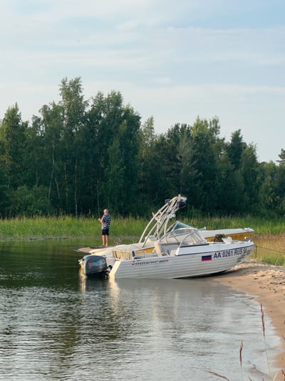 In Borovoye, Russia, a man stands on a wooden pier, fishing with a rod. He is dressed in a patterned shirt and dark shorts. A white motorboat with a wakeboard tower is beached on a sandy shore to the right, its registration number "AA 0261 RUS 54" visible. Another yellow boat is positioned behind it. The scene is set against a backdrop of lush green trees and a calm, reflective body of water. The sky above is a pale blue with wispy clouds. The overall impression is one of a peaceful outdoor recreation setting.