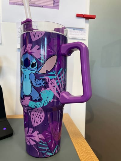 An extreme close-up shot of a purple travel tumbler featuring the Disney character Stitch is presented. The tumbler has a floral pattern with large leaves and flowers in shades of purple, pink, and teal. Stitch is depicted in his classic blue color, with large ears and a mischievous grin, positioned on the lower half of the tumbler. A purple handle is attached to the right side of the tumbler, and a white straw with a purple silicone tip protrudes from the top. The tumbler is placed on a gray surface, possibly a desk, with a wooden surface in the background to the right. The background is blurred, but hints of white and dark gray walls are visible, along with a glimpse of a black laptop on the left. A red clip is visible on the white wall behind the tumbler. The lighting suggests an indoor environment, possibly an office or home setting. There is no identifiable information or context that would place this image in Morsbach, Germany.