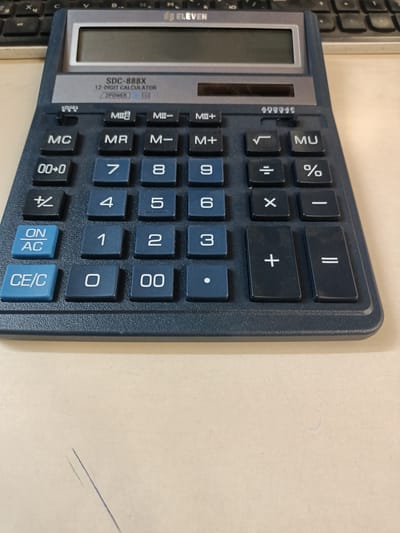 This is a close-up, eye-level, vertical shot of a dark blue SDC-888X 12-digit calculator with a dual power source, placed on a light-colored surface, likely a desk. The calculator has a silver top panel with the brand "ELEVEN" printed on it, and a digital display screen. Below the display are various buttons, including memory functions (MC, MR, M-, M+, MU), arithmetic operations (+, -, x, ÷, =, %), number keys (0-9, 00), and control keys (ON/AC, CE/C). The calculator appears to be in use or ready for use, with no specific activity or event taking place other than its presence.

Notable details include the text "SDC-888X 12-DIGIT CALCULATOR 2POWER" and a serial number "A03345" visible on the calculator. The background shows a hint of a computer keyboard, suggesting the calculator is in an office or study environment. The lighting is even, indicating it's likely indoors, possibly during daylight hours. The surface beneath the calculator shows some minor scratches and smudges, suggesting it's a well-used item or workspace. There are no people or animals in the frame. The context provided suggests the location is Mikhnevo, Russia, but this information is not visually discernible from the image itself.