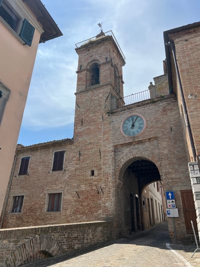 The image shows an upward view of a brick tower with a clock and a narrow street lined with brick buildings. The tower has a large, round clock face with Roman numerals, and a bell tower structure at its top. Below the clock, there is an arched passageway leading into a darker area. The street is paved and has a white line painted on it, suggesting it might be a road or pathway. Several windows with dark shutters are visible on the brick buildings. Signage is present above the archway, including a blue sign with white arrows indicating direction and parking symbols, and another sign with a red cross, possibly indicating a medical facility. The sky is blue with some clouds. The architecture suggests a historic European town, consistent with the provided location context of Barbara, Italy. The lighting indicates it is daytime, likely late morning or early afternoon given the shadows. There are no people visible in the image.