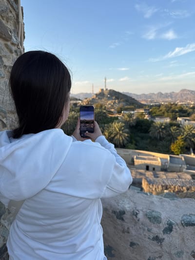 A female individual, seen from the back, stands on an elevated stone wall overlooking a landscape in Hatta, United Arab Emirates. She has dark hair and wears a white hooded sweatshirt. The individual holds a black smartphone horizontally with both hands, aiming it towards the distant panorama, with the phone's screen displaying the real-time captured view. The background features rugged mountains under a blue sky with scattered white clouds. The mid-ground contains numerous palm trees and traditional, light-colored buildings with thatched roofs. A prominent fort-like structure or tower, topped with a communication mast, is visible on a distant rocky hill. The scene is illuminated by natural daylight.