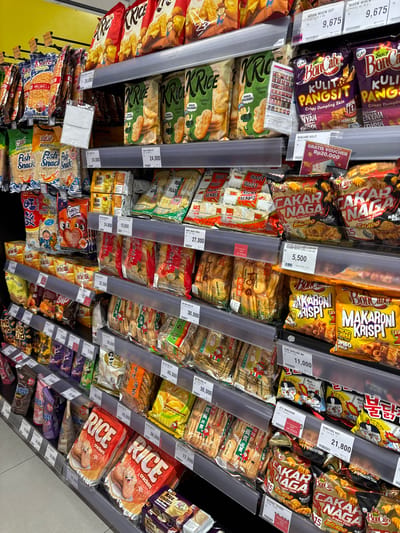 The image captures a tilted view of a snack aisle in a supermarket or grocery store in Kelapa Dua, Indonesia. The shelves are fully stocked with a variety of packaged snack foods, primarily chips, crackers, and crisps. No people are visible in the frame.

Numerous brands and products are identifiable on the colorful packaging. Prominently displayed are "Boncabe Kulit Pangsit" (Crispy Dumpling Skin), "Kriuk Rice" with seaweed flavor, "Cakar Naga" (Dragon Claw) snacks in various flavors like Balado, "Makaron Krispi" (Crispy Macaroni) with nacho cheese flavor, and "Samyang Hot" spicy noodles. Other visible products include "Jetz Makaroni," "Want-Want Senbei," "Fish Snack" (ikan) products, and multiple types of "Rice Crackers" (including "Bin Bin Rice Crackers" and "O'Rion K'R").

Price tags are affixed to the shelves below many products, indicating prices in Indonesian Rupiah (Rp). Examples include Rp 9,675 for Miow Miow Cut, Rp 5,500 for Kobe Boncabe Cakar, Rp 11,000 for Kobe Boncabe Ma, Rp 21,800 for Samyang Hot, Rp 24,000 for Delfi Orion K'R, Rp 27,000 for Want Want Shell, and prices ranging from Rp 12,800 to Rp 45,500 for other items. A promotional sign offering a "GRATIS VOUCHER Rp20.000" (Free Voucher Rp20,000) for Boncabe Kulit Pangsit is visible. There are also "OUT OF STOCK" labels on some shelf sections.

The setting is brightly lit, consistent with an indoor retail environment. The wide array of snack options suggests a diverse selection catering to different tastes.