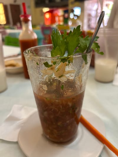 The image captures a prawn cocktail in a glass, garnished with celery. A spoon is seen in the glass. The drink rests on a white plate, alongside a carrot stick. In the background, a bottle of sauce is visible. The location is Veracruz City, Mexico, judging by the presence of the prawn cocktail. The scene appears to be indoors, possibly a restaurant.