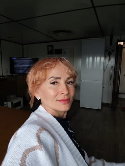 A medium shot shows a woman with short, peachy-pink hair and fair skin posing for a selfie. She is wearing a white knit cardigan with a tan geometric pattern over a dark blue top with a white abstract design. She has on makeup, including eyeliner and pink lipstick, and is wearing a small, heart-shaped earring with rhinestones in her left ear.
The background suggests an interior setting, possibly a residential or communal space. To the left, a dark wooden table is visible, and behind that, a black television screen displays a blurred image. Further back, a white refrigerator stands against a wall, and to its right, a doorway leads to another room. The ceiling is dark and appears to be constructed with paneling and beams. The floor is made of dark wood planks. The lighting is dim, with some light emanating from the doorway. The overall impression is of a casual, indoor snapshot. The provided image and its crops do not contain any information that would indicate the location is the city of Ozernoe Ust'e, Russia.