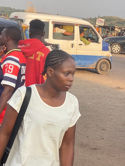 A woman in a white top looks off to the side. Braided hair is visible. Behind her, three people are wearing colorful shirts. A white van with a blue stripe and an advertisement is adjacent. The van is open, with two passengers inside. The street is visible.