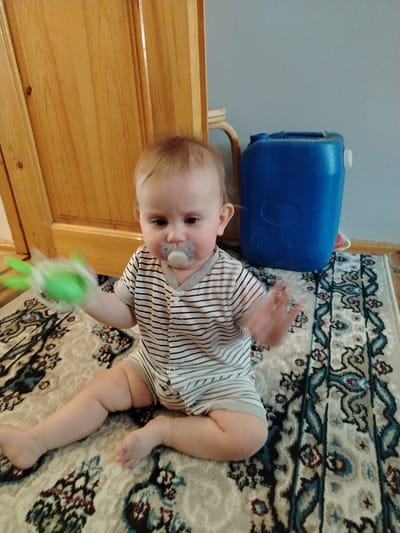 An infant is sitting on a rug in what appears to be a room, likely in Shovot, Uzbekistan. The infant is wearing a striped onesie and has a pacifier in its mouth. In its right hand, the infant is holding a green toy. To the right of the infant, there is a large blue plastic container. Behind the infant is a wooden door. The floor is covered with a patterned rug. The lighting suggests an indoor environment.
