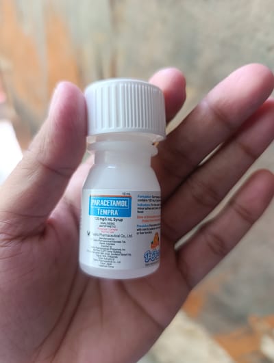 A close-up shot shows a person's hands holding a small white plastic bottle of "PARACETAMOL TEMPRA" syrup. The bottle has a white ribbed cap and a blue and white label. The label indicates "10 mL", "120 mg/5 mL Syrup", "ANALGESIC", "ANTIPYRETIC", and "Physician's Sample Not For Sale". It also mentions "Manufactured by: PT Taisho Pharmaceutical Indonesia Tbk. Depok, Indonesia" and "Imported by: Taisho Pharmaceuticals (Philippines), Inc. 16th Floor CUIPP Center Building, 11th Avenue corner 35th Street, Bonifacio Global City, Taguig City 1634, Philippines". The background is blurred, showing an orange and beige textured surface, possibly a wall or floor. There is no visual information in the image to determine the specific location within the city of Kawit, Philippines.