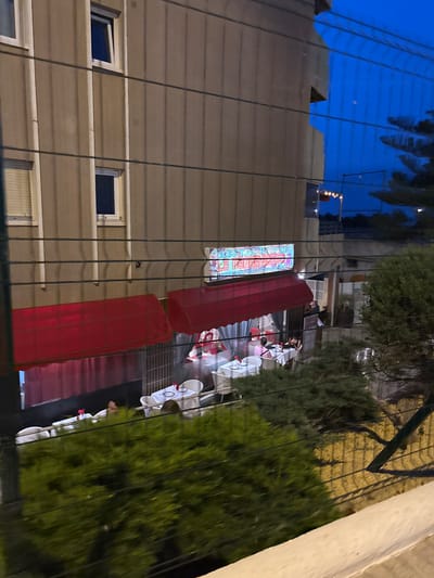 In Calp, Spain, a restaurant with red awnings is visible at dusk. The restaurant's name, "RESTAURANTE LE INDIENNE," is displayed on a neon sign. Several tables with white tablecloths are set outside, some with diners. The building appears to be an apartment complex, with windows visible on the upper floors. A fence runs across the foreground, partially obscuring the view. Lush green foliage is present in the lower portion of the image. The sky is a deep blue, indicating evening.