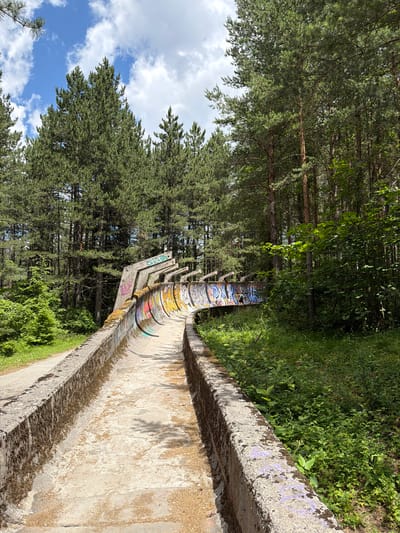 This image shows the abandoned bobsled track from the 1984 Winter Olympics, located on Mount Trebević in Sarajevo, Bosnia and Herzegovina. The concrete track, worn and covered in moss and graffiti, winds through a dense forest of tall pine trees. The sky above is blue with scattered white clouds. The track's concrete walls are covered in colorful, graffiti art. Vegetation, including grass and bushes, grows around the track and on its sides. A narrow paved path runs alongside the track on the left side of the image. The scene conveys a sense of decay and abandonment within a natural setting.