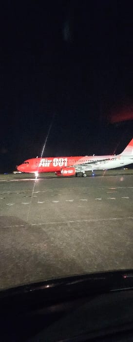 A vertical photograph taken at night depicts an "Air 001" branded Airbus A320 series aircraft stationary on an asphalt taxiway at an airport in Pyrzowice, Poland. The aircraft's fuselage is primarily red, featuring "Air 001" in large white text on its side. Its engines also display "Air" branding in white. The tail section is predominantly red, transitioning to white at the upper rear edge. Bright landing or taxi lights emanate from the front of the aircraft, illuminating the tarmac directly ahead. The ground surface consists of asphalt with visible white dashed and solid markings. The sky overhead is dark, indicating night. The image is framed from a low perspective, with the dark upper edge of a vehicle's dashboard or hood visible at the bottom of the frame. No individuals, specific ground support equipment, or active interactions are discernible.