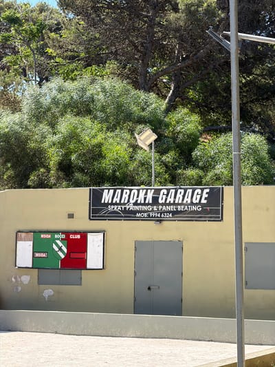The image shows a light yellow building facade in Pietà, Malta, with a black banner that reads "MAROKK GARAGE SPRAY PAINTING & PANEL BEATING MOB: 9994 6324". Below the banner, a score board with "MSIDA BOCCI CLUB" and what appears to be bocce ball imagery is mounted on the wall. To the right of the scoreboard is a grey metal door with a padlock, and to its right, a grey shuttered window. A metal pole with an industrial light fixture is visible on the right side of the image. Dense green foliage and trees are behind the building. The ground in front of the building is sandy or gravelly, bordered by a low concrete wall.