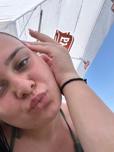 A close-up, selfie-style shot captures a young woman with her hand near her face, holding a lit cigarette. She is wearing a green bikini top and a black bracelet with green accents on her wrist. Her lips are pursed, and her skin appears wet, suggesting she may have been swimming. The background shows a white umbrella with red logos and a clear blue sky, consistent with an outdoor setting, possibly a beach or pool area in Manolsko Konare, Bulgaria. The lighting is bright and natural, indicative of daylight.