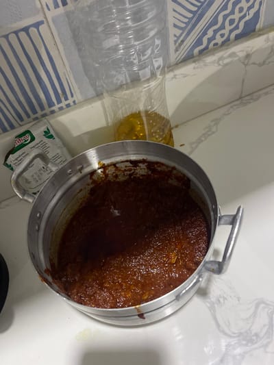 The image displays a close-up indoor scene, likely in a kitchen or food preparation area. The primary focus is a metallic pot, silver in color, tilted on its side, revealing a significant amount of dark reddish-brown, thick liquid or paste-like substance inside. This substance has a somewhat textured, lumpy appearance, consistent with a stew, curry, or sauce. The pot has two small, metallic handles on opposite sides.
To the left of the pot, a clear plastic bottle is visible. The bottle is partially filled with a golden yellow liquid, which appears to be cooking oil. The top part of the bottle seems to be cut or modified, suggesting it might be used for pouring or as a makeshift container. The bottle is lying horizontally on a surface.
Below the bottle and partially obscured by the pot, there is a small white packet with green text. Visible text on the packet includes "Dew", "Ingredients:", and "Sugar Free". Further down, the text "Ingredients: Veggies, Potato" can be partially deciphered, suggesting it might be a seasoning mix or an instant food packet.
The surfaces in the image consist of different materials. To the left, there's a countertop or wall with blue and white striped tiles arranged in a wavy pattern. Above this, and extending behind the pot, is a white surface with subtle grey marble-like patterns, possibly a countertop or backsplash. There are a few small splashes or stains of the reddish-brown substance from the pot on the white surface near the pot.
The lighting suggests an indoor setting, with no direct indication of the time of day. The scene captures items typically found in a cooking environment, with ingredients (oil, possibly seasoning) and a prepared dish in a pot.