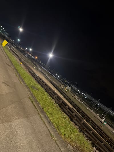The image shows a view of railroad tracks at night, likely in Dudelange, Luxembourg based on the location request. The tracks run diagonally across the frame, illuminated by distant streetlights. To the right of the tracks, there's a narrow strip of grass and a concrete surface, possibly a sidewalk or platform. The lighting is dim and the sky is dark, indicating it's nighttime. There's a yellow container near the lower left of the frame. The perspective is tilted slightly to the left, adding a dynamic feel to the scene.
