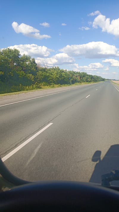 The image shows a view from inside a vehicle looking out at a highway. The road is paved with asphalt and has a white line marking the lane. On the left side of the road, there is a strip of vegetation, followed by a line of trees. The sky is blue with scattered white clouds. In the distance, a few cars can be seen on the highway. The foreground shows the dashboard and a shadow of the vehicle's side mirror. The scene appears to be a sunny day on a highway in a rural or suburban area. The location is identified as Золотая Степь, Russia.