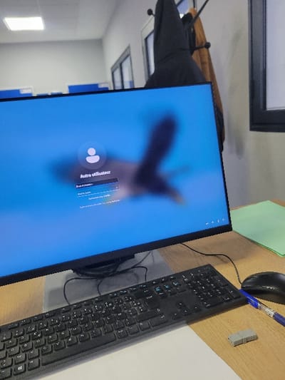 A desktop setup features a monitor displaying a login screen with a user icon and text in French, a black keyboard, a mouse, a blue pen, and a small USB drive on a wooden-like surface. The background includes a glimpse of a building interior with windows, a coat rack with jackets, and a green document partially visible. The monitor is angled towards the right side of the image, with the keyboard aligned closely beside it.