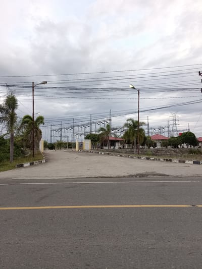 This media file depicts an overcast day at an electrical substation in Dayah Teungoh, Indonesia. The scene is dominated by an array of electrical towers, transformers, and overhead power lines stretching across the frame. In the foreground, a paved road with a yellow dividing line runs horizontally, indicating a public thoroughfare. Adjacent to the road, a paved area leads towards the substation entrance, bordered by grass and small trees.
Several palm trees are visible, adding a tropical element to the landscape. Beyond the entrance, small buildings with red roofs are situated within the substation compound. The sky is filled with heavy, gray clouds, suggesting a cloudy or possibly rainy day. There is no visible activity or people in the image, creating a sense of stillness. The overall impression is one of industrial infrastructure set against a natural, albeit subdued, environment. The time of day is difficult to determine precisely due to the overcast conditions, but it appears to be daytime. There is no visible text in the image.