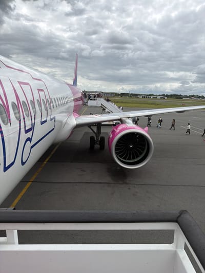 This image captures an airport tarmac scene from an elevated viewpoint, likely from the top of an air stair or boarding bridge. A large Wizz Air passenger jet, identifiable by its distinctive white, pink, and blue livery, dominates the foreground. The stylized "Wizz Air" branding is clearly visible in pink and blue along the fuselage, which also features numerous passenger windows.

The aircraft's right wing, extending towards the viewer, houses a prominent engine with a pink cowling and visible fan blades. Several people are scattered across the grey asphalt tarmac, walking towards or away from the aircraft's front entrance, indicating ongoing boarding or disembarkation.

The sky is heavily overcast with dense grey and white clouds, suggesting a cloudy daytime setting. In the distance, glimpses of airport buildings and a green landscape are visible. The immediate foreground shows the white metal railings and structure of the boarding ramp. The tarmac features typical yellow operational markings. The event is clearly an active airport operation, with passengers and an aircraft present at Tillé, France.