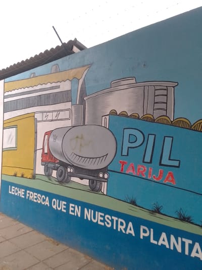 The image shows a mural painted on a wall. The mural depicts a milk factory with a truck delivering milk. The truck is gray and has a smiley face on its tank. To the right of the truck, the word "PIL" is painted in blue, with "Tarija" in red underneath. Below the truck, the text "LECHE FRESCA QUE EN NUESTRA PLANTA" is written in white. The location is Municipio Tarija, Bolivia, as indicated by the text on the mural.
