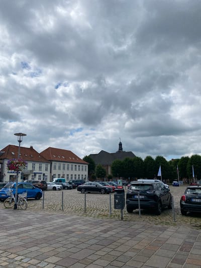 The image captures a view of the Obereider promenade in Rendsburg, Germany, on a cloudy day. The foreground is dominated by a paved plaza with a distinctive pattern of square and rectangular stones. A light-colored bicycle is parked on the left side of the plaza, leaning against a post. Several cars are parked in the background, including a blue hatchback, a white sedan, a black SUV, and a black Mini Cooper.

Behind the cars, a row of buildings lines the plaza. The buildings have traditional red-tiled roofs and white facades with numerous windows. One building on the left appears to house a "Garnison" store and a pharmacy. Further back, a large, dark-colored church building with a prominent tower stands amongst a line of lush green trees. Several flags are visible on poles in front of the trees. The sky is overcast with heavy, grey clouds, interspersed with patches of blue.