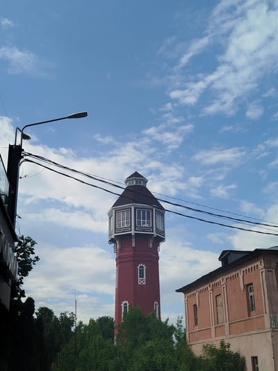 The image shows a water tower in Noginsk, Russia. The tower is made of red brick and has a polygonal top section with windows. A light pole and electrical wires are visible in the foreground. Trees are visible in the middle ground, partially obscuring the base of the tower. An old building is located to the right of the tower, constructed of pink brick with ornate designs. The sky is blue with scattered white clouds.