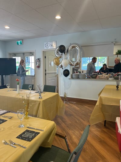 Here is a detailed description of the image:
**General Description:**
The image depicts an interior space prepared for a celebration, likely a 90th birthday party. The space is well-lit and features tables set up for guests, along with individuals preparing food in the background.
**Location:**
The clock on the wall shows "Tarkett" written on it. This indicates the image may have been taken in Kelowna, Canada.
**People:**
* There are several individuals visible in the background, behind a counter area. They appear to be involved in food preparation or serving.
* A woman is standing near the entrance, holding a smartphone, possibly taking a picture or video.
**Subjects:**
* The primary subject is the setup for a 90th birthday celebration.
**Objects:**
* Large "90" balloons in silver and gold.
* Black, gold and white balloons with phrases like "Happy Birthday To You" printed on them.
* Tables draped in gold tablecloths, set with silverware, glassware, and table number markers (e.g., "06").
* Floral centerpieces and other decorative elements on the tables.
* A serving counter with food items and plates.
* A clock on the wall.
* A television in the corner.
* An exit sign.
**Actions and Interactions:**
* The individuals behind the counter are engaged in food preparation or serving.
* The woman is using her smartphone.
**Environment:**
* The interior has wooden flooring, light-colored walls, and a drop-ceiling with recessed lighting.
* There are windows that provide natural light.
* The space appears to be a community hall or event room, likely designed for gatherings.
This describes the scene based on the visual information available in the image.